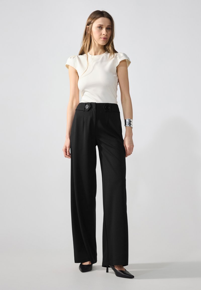Pantaloni neri a gamba larga con due bottoni frontali, abbinati a un top aderente crema con maniche a sbuffo, indossati con tacchi neri a punta.