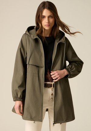 Kobieta w oliwkowozielonej kurtce z kapturem, czarnym topie i beżowych spodniach, pozująca na jednolitym beżowym tle.