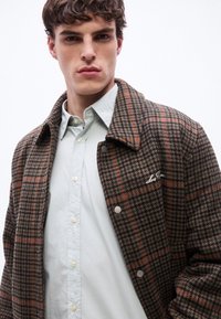 Νεαρός άνδρας που φορά καφέ και πορτοκαλί καρό σακάκι πάνω από ανοιχτογκρι πουκάμισο με κουμπιά, κοιτάζοντας μπροστά με ουδέτερη έκφραση.