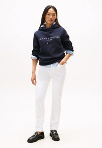 Nő áll, sötétkék Tommy Hilfiger kapucnis pulóvert, világoskék inget, fehér nadrágot és fekete mokaszint viselve, egyszínű fehér háttér előtt.