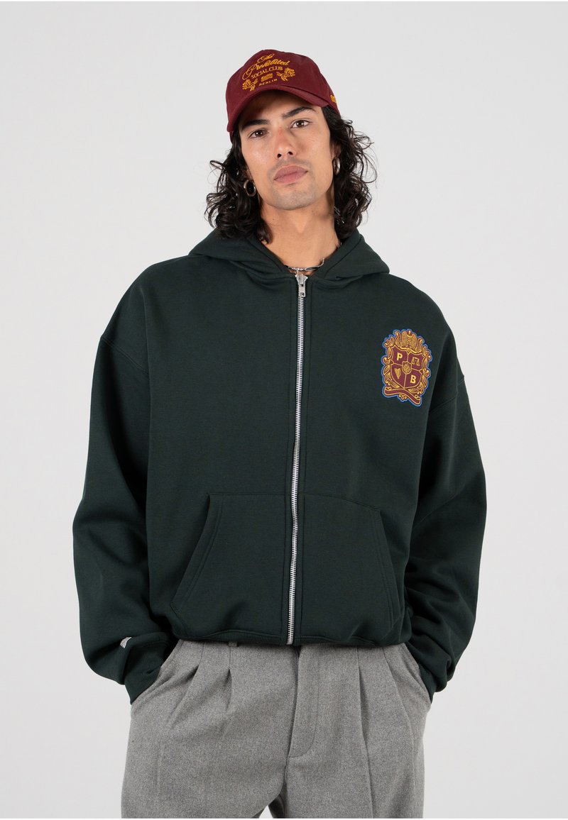 Felpa zip grigia scura con tasca frontale e patch ricamato. Abbinata a un cappellino bordeaux con testo giallo. Pantaloni grigi con pieghe.