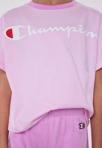 Camiseta de algodón rosa con cuello redondo y mangas cortas, que presenta un gran logo blanco de "Champion" y un acento rojo. Combinada con pantalones rosas.