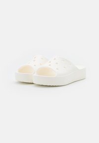 Vita slip-on sandaler med upphöjda sulor och flera ventilationshål på toppen, visade mot en neutral ljus bakgrund.