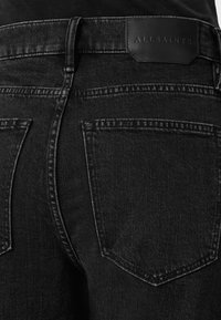 Zwarte denim jeans met een gestructureerde afwerking, voorzien van achterzakken en een leren logo patch op de tailleband. Gedetailleerde stiksels zichtbaar.