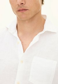 Camicia bianca in lino con colletto, bottoni frontali e una tasca sul petto. Il tessuto è testurizzato e leggero.