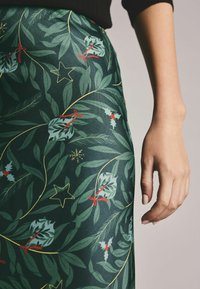 Jupe vert foncé ornée d'un motif de feuilles, de houx et d'étoiles en rouge et or. Texture lisse avec une coupe ajustée. Main visible sur le côté.