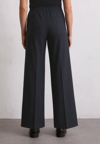 Pantalons à jambes larges en tissu noir uni, dotés d'une taille élastique et d'une discrète poche arrière, sur un fond neutre.