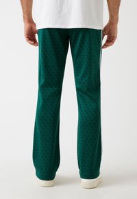 Pantaloni verdi a fantasia realizzati in un tessuto morbido, caratterizzati da una forma svasata e strisce laterali. Sneakers bianche visibili alle caviglie.