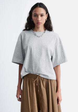 Kvinde med langt mørkt hår iført en grå oversize t-shirt, brune bukser med snøre, sølvkæde, creoløreringe og ringe.