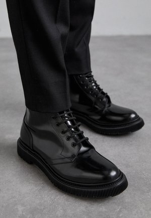 Svarta läderankleboots med en slät yta, snörning och ribbad svart gummisula. Bärs med mörka byxor på en grå yta.