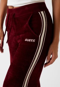 Bordové velúrové teplákové nohavice s bielymi bočnými pruhmi. Obsahujú nastaviteľný pás so šnúrkou a malé logo "GUESS" na ľavom stehne. Mäkká textúra.