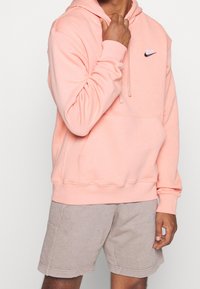 Sudadera con capucha de color rosa claro, con un bolsillo canguro y un pequeño logo negro de Nike en el pecho. Presenta puños y dobladillo acanalados.