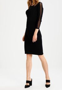 Robe noire avec de longues manches transparentes, texture côtelée, silhouette ajustée, longueur au genou, portée avec des chaussures à talons hauts noires.