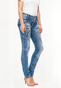Jeans skinny in denim blu chiaro con dettagli sbiaditi, numerose tasche e cuciture a contrasto. Indossati con un top aderente bianco e tacchi beige.