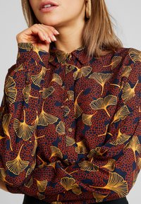 Chemise à manches longues en tissu bleu foncé ornée d'un motif vif de feuilles de ginkgo rouges et dorées, avec boutonnage sur le devant et col classique.