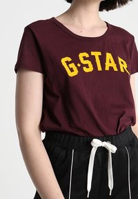 Camiseta burdeos con texto amarillo "G.STAR", de mangas cortas y escote redondo. Combinada con pantalones negros que tienen cordón blanco y detalles laterales.