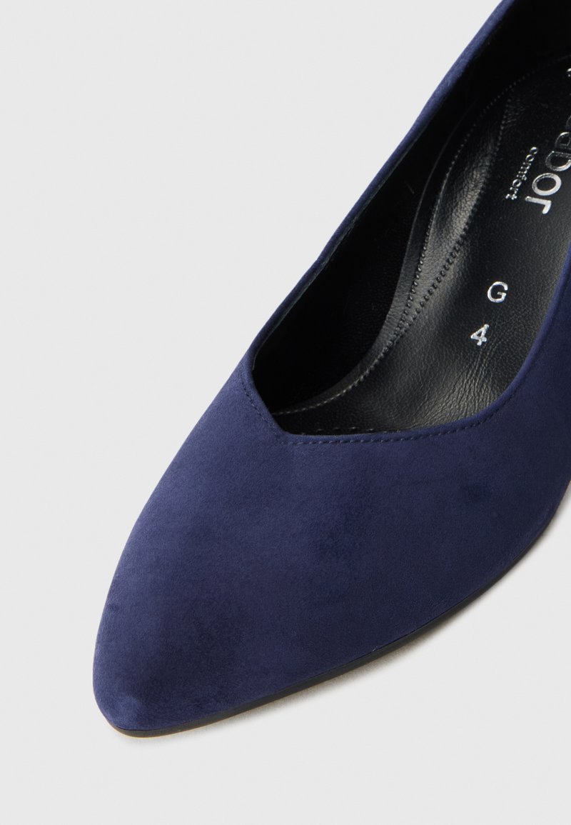 Chaussure à talon haut en daim bleu foncé avec un bout pointu et un design découpé subtil, posée sur un fond gris clair.