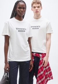 Λευκά βαμβακερά T-shirt με την επιγραφή «TOMMY JEANS» σε μαύρο, συνδυασμένα με σκουρόχρωμα τζιν παντελόνια. Ένα μοντέλο κρατάει μαύρη τσάντα χειρός, ενώ το άλλο έχει ένα κόκκινο καρό πουκάμισο δεμένο στη μέση.