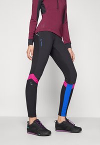 Fekete sportos leggings rózsaszín és kék díszítéssel, testhezálló fazonnal és rugalmas anyagból készült. Fekete sportcipővel párosítva.