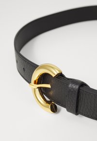 Ceinture en cuir noir avec une grande boucle circulaire en or présentant un design découpé distinctif. Surface texturée avec des coutures visibles et des trous.