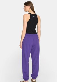 Kobieta w czarnej bezrękawnikowej bluzce i luźnych fioletowych spodniach, stojąca z boku, z głową lekko odwróconą w tył.