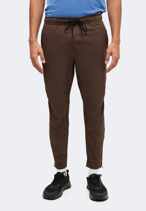 Homme portant un pantalon marron fuselé avec une taille à cordon, des chaussettes blanches et des chaussures de sport noires sur un fond clair.