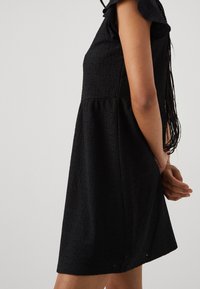 Robe noire avec des manches courtes à volants, un tissu à texture optique avec des motifs à œillets, et une silhouette évasée qui tombe au-dessus du genou.