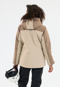 Beige og brun isoleret jakke med hætte, der har en glat tekstur og subtil farveblokeringsdesign. En hånd holder en hvid hjelm.