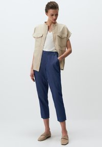 Beżowa koszula z krótkim rękawem z dwoma kieszeniami na piersi, biała bluzka z dekoltem w serek oraz niebieskie spodenki. Beżowe buty wsuwane dopełniają całość.