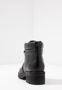Zwarte leren enkelboot met chunky hak, gewatteerde kraag en metalen veterogen, weergegeven van achteren op een witte achtergrond.