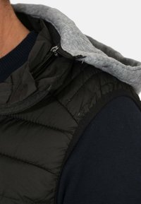 Gilet invernale nero con texture trapuntata, chiusura con zip e fodera in felpa grigia. Presenta un tessuto a maglia blu navy sulle spalle.