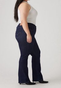 Jean évasé bleu foncé en denim, doté d'une taille haute, de poches arrière et de détails de couture visibles. Porté avec un débardeur blanc.