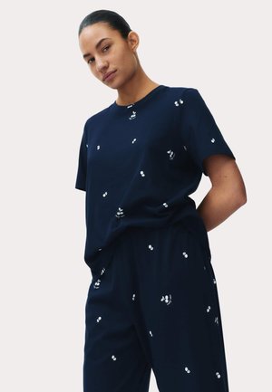 Vrouw die een marineblauwe top met korte mouwen en een broek draagt met kleine witte kersenpatronen, staand met haar handen achter haar rug tegen een lichte achtergrond.