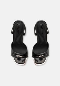 Black patent leather slingback heels with a square toe, featuring a metallic accent and embossed logo on the strap. Smooth texture.