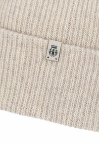 Helle beige Strickmütze aus weicher Wolle, mit einem gerippten Textur und einem kleinen Stoffetikett mit einem Logo und einem komplexen Design.