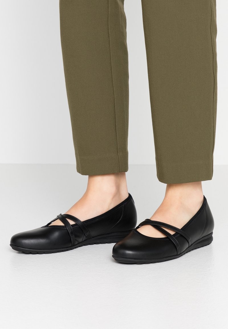 Pieds portant des ballerines en cuir noir à brides croisées, associées à un pantalon court vert olive, debout sur une surface lisse.