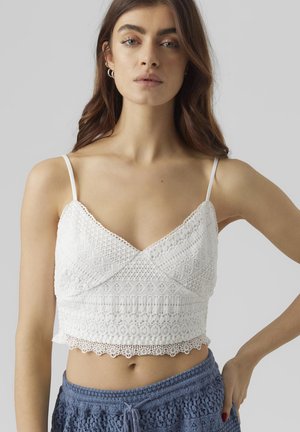 White cropped top made of intricate lace, featuring adjustable spaghetti straps and a scalloped hem. Textured pattern throughout the fabric.