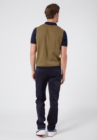 Knitted vest with a yellow and navy geometric pattern, paired with dark trousers and white sneakers. Visible ribbed collar and hem details.