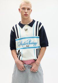 Gestreiftes Poloshirt in Creme, Marineblau und Blau mit kurzen Ärmeln und einem knöpfbaren Kragen. Mit dem Text "Miglio Punteggio" in Kursivschrift. Graue Hose.