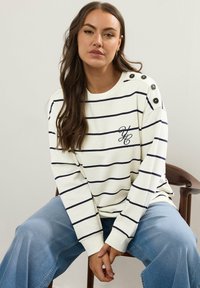 Vit långärmad topp med marinblå ränder, med knappdetaljer på axlarna och en broderad logotyp. Parat med blå vida jeans.