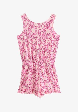 Ung pige står og smiler i ærmeløs lyserød blomstret romper med flæsekant, hvide sneakers og matchende lyserød scrunchie på håndleddet.