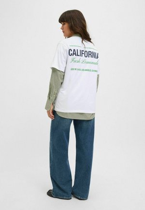 Bílý tričko s zeleným nápisem "CALIFORNIA Fresh Lemonade" na zadní straně. Pod ním pruhovaný zelený dlouhý rukáv a široké modré džíny.
