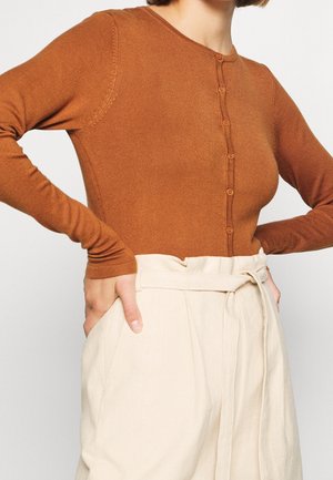 Vrouw die een nauwsluitend bruin topje met lange mouwen en knopen draagt, ingestopt in een hooggetailleerde crèmekleurige paperbagbroek, met handen op de heupen.