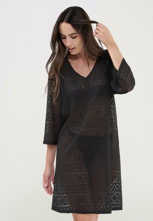 Mujer con cabello largo y castaño, vestida con un vestido negro semitransparente con patrones geométricos, mirando hacia abajo y tocándose el cabello.