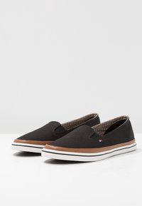 Un paio di scarpe slip-on in canvas nere con bordi marroni e suole bianche, caratterizzate da un piccolo logo con bandiera rossa, bianca e blu sul lato.