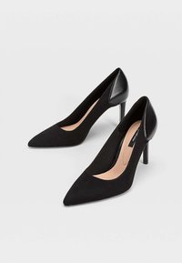 Zapatos de tacón negro con diseño de punta afilada, hechos de un tejido texturizado, que presentan un acento de tacón de cuero negro liso y un forro interior neutral.