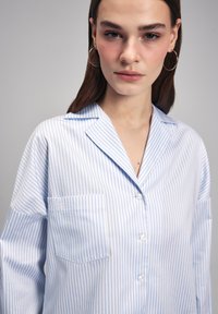 Camicia a righe blu chiaro e bianca, con taschino sul petto, morbida al tatto e dal taglio rilassato. Presenta un colletto classico e maniche lunghe.