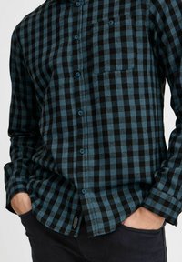Camicia in cotone a quadri in teal e nero con maniche lunghe, bottoni blu, tasca sul lato sinistro del petto e orlo curvato. Presenta un motivo a scacchi.