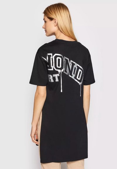 Donna con capelli biondi che indossa una maglietta nera oversize con lettere argentate che colano sul retro, abbinata a pantaloni beige.