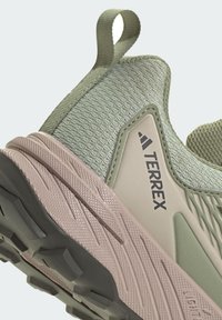 Lichtgewicht groene sportschoen met een gebreide bovenkant, versterkte neus en een roze, gestructureerde buitenzool met een opvallend profielpatroon.